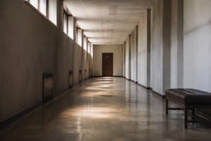Empty corridor in an older civic building with late afternoon light on worn flooring and a single closed office door at the far end.