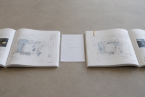 Two identical plan documents open side by side on a desk at visibly different pages, each annotated by a different hand in pencil.